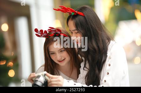 Due giovani donne che indossano cerchietti festosi con corna di renne si deliziano nello spirito natalizio mentre guardano le foto con una macchina fotografica. I loro sorrisi Foto Stock