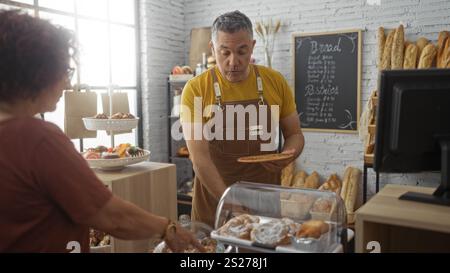 Man baker, che serve una cliente donna all'interno di un panificio, presenta una selezione di pasticcini e pane in un ambiente rustico. Foto Stock