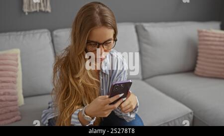 Giovane donna con capelli biondi e occhiali che usa lo smartphone in un accogliente soggiorno con divano grigio Foto Stock