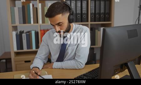 Uomo ispanico che indossa le cuffie e scrive alla sua scrivania in un ufficio pieno di libri e file. Foto Stock