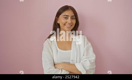 Una donna in piedi con le braccia incrociate su uno sfondo rosa mette in mostra bellezza e sicurezza, mettendo in evidenza le sue giovani caratteristiche ispaniche in un isolato st Foto Stock