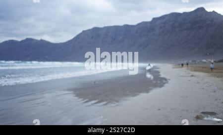 Vista sfocata di una spiaggia sabbiosa con montagne sullo sfondo e persone che passeggiano vicino al litorale sotto un cielo nuvoloso Foto Stock