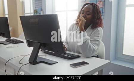 Donna afroamericana che lavora al chiuso in una stanza d'ufficio, guardando un computer mentre guarda premurosa. Foto Stock