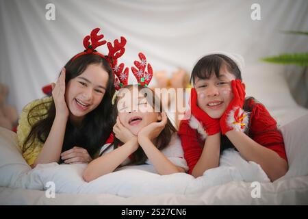 Tre bambini festeggiano il Natale, indossando corna di renne e accessori a tema Babbo Natale. Stanno posando giocosamente su un letto Foto Stock