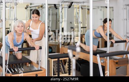 Donna anziana positiva che fa esercizi sul riformatore in studio pilates con una giovane personal trainer che controlla i suoi movimenti. Foto Stock