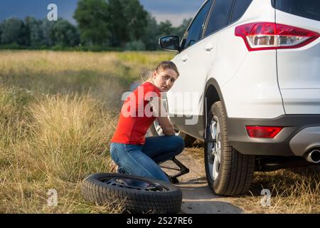 Donna seduta ad un'auto rotta e che cerca di cambiare la ruota forata Foto Stock