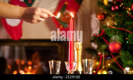 Immagine ravvicinata della mano che accende la candela rossa contro l'albero di Natale e il camino Foto Stock