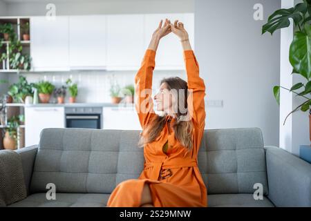 La donna si allunga felicemente con le mani alzate e si rilassa sul divano nel soggiorno. Weekend pigri, disintossicazione digitale Foto Stock