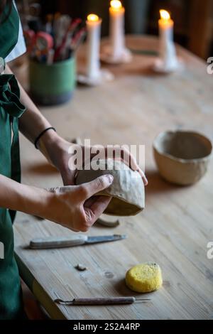 Le mani di donna potter modellano una ciotola di argilla bianca su un tavolo illuminato da candele di legno nello studio di laboratorio di ceramica Foto Stock