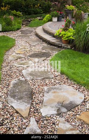 Sentiero in pietra e ghiaia attraverso prato verde curato che conduce al patio in pietra con pavimentazione marrone rialzata con piantatrici nel giardino residenziale sul retro. Foto Stock