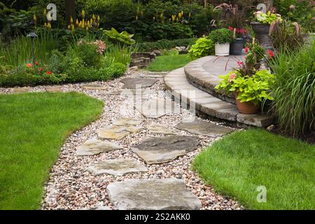 Sentiero in pietra e ghiaia attraverso prato verde curato che conduce al patio in pietra con pavimentazione marrone rialzata con piantatrici nel giardino residenziale sul retro. Foto Stock