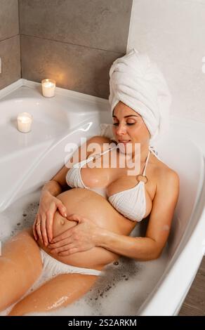 Donna incinta che gode di un bagno di bolle con candele Descrizione Foto Stock