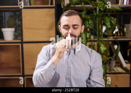 Il giovane si siede al lavoro e usa una bottiglia di spray nasale con spazio negativo per inserire il testo. Foto con messa a fuoco selettiva. Foto Stock