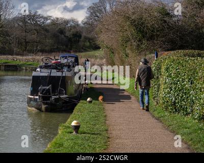 Passeggia per cani su un sentiero lungo il canale, passando davanti a una barca ormeggiata in una soleggiata giornata invernale, Stoke Bruene, Northamptonshire, Regno Unito Foto Stock
