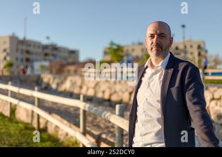 Un uomo professionista con un abito sartoriale si trova accanto a una recinzione in legno rustica, con un cielo azzurro. Ha una posizione sicura e un abbigliamento affilato Foto Stock