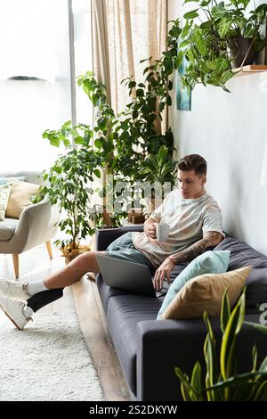 Il giovane uomo si diverte a stare sul divano, a lavorare su un computer portatile sorseggiando un caffè. Foto Stock