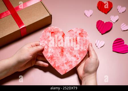 Donna che tiene in mano un biglietto d'auguri fatto a mano a forma di cuore per la giornata di San Valentino. Copiare lo spazio su sfondo rosa. Cuori rossi di san valentino. Disposizione piatta, parte superiore vi Foto Stock