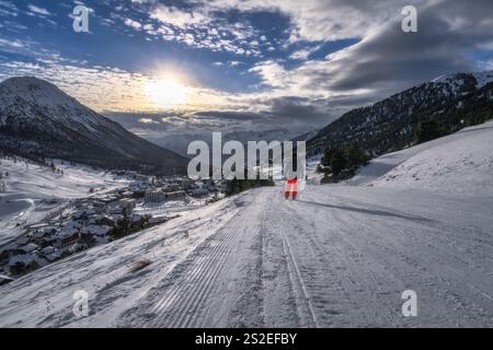 Un tranquillo paesaggio invernale presenta uno sciatore solitario che scivola su un sentiero innevato, incorniciato da montagne e da un tramonto mozzafiato che diffonde calde sfumature dorate, Foto Stock