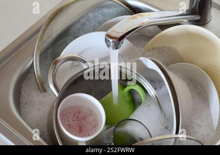 I piatti sporchi e gli elettrodomestici da cucina non lavati si trovano nell'acqua di schiuma sotto un rubinetto da un rubinetto della cucina Foto Stock