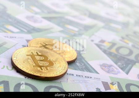 Le bitcoins fisiche dorate si trovano su un insieme di tagli monetari verdi di 100 euro. Un sacco di soldi forma un mucchio infinito Foto Stock