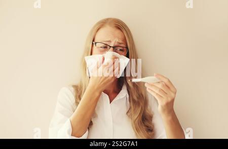 Malato turbato dipendente donna caucasica starnuti colpo naso utilizzando il tessuto e misurare la temperatura corporea guardando il termometro a casa Foto Stock