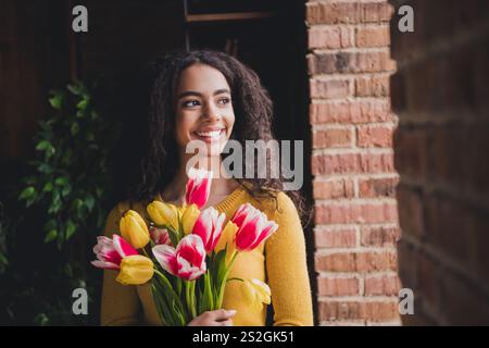 Giovane donna che tiene in mano tulipani colorati mentre sorride vicino a un muro di mattoni in interni accoglienti Foto Stock