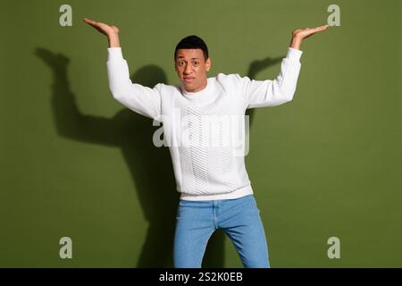 Il giovane e affascinante uomo in un maglione bianco casual posa giocosamente con le mani alzate sullo sfondo cachi Foto Stock