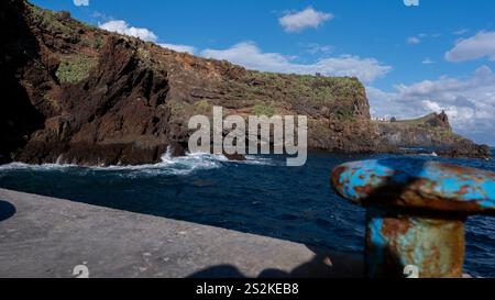 Una scena costiera con aspre scogliere ricoperte di vegetazione, onde che si infrangono sulle rocce e un ormeggio arrugginito in primo piano sotto un cielo soleggiato. Foto Stock