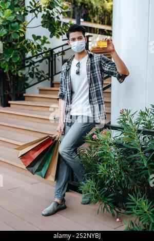 Ritrattistica giovane uomo in maschera con borsa di carta e carta di credito sulle scale del centro commerciale, nuovo stile di vita normale e concetto di shopping Foto Stock