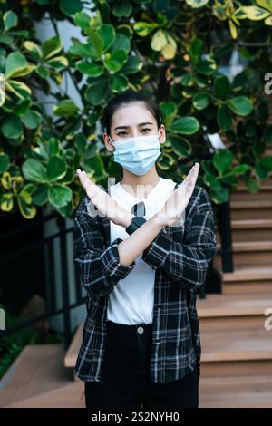 La bella giovane donna con la maschera di protezione mostra la croce con il segno della mano sulle scale di un centro commerciale, New Normal Lifestyle e shopping Foto Stock