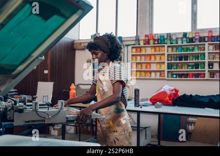 Vista del profilo di un operatore grafico multiculturale sorridente in piedi vicino alla pressa per serigrafia e con un piatto di stampa. Giovane professionista fem Foto Stock