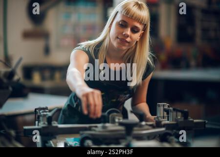 Lavoratrice professionista dell'industria grafica sorridente in piedi presso l'officina di stampa e che opera la macchina di stampa serigrafica. Un'officina di stampa esperta Foto Stock