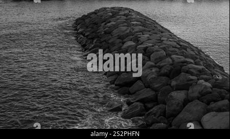 Immagine in bianco e nero di una scogliera rocciosa che si estende fino al mare, circondata da acque calme e lievi onde vicino alla riva. Foto Stock