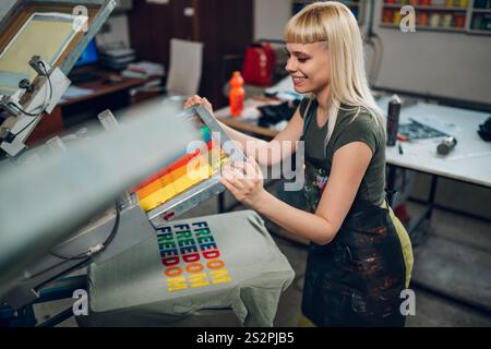 Una donna sorridente, un tecnico grafico esperto, esegue la stampa serigrafica su una macchina di stampa manuale a carosello presso l'officina di stampa. Stampa professionale Foto Stock