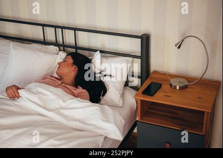 La mattina, la giovane e bellissima donna sdraiata nel suo letto e che dorme con il suo smartphone sul comodino Foto Stock