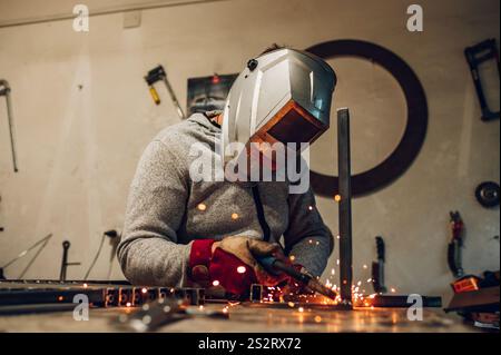 Lavoratore in fabbrica che salda metallo con saldatrice mentre indossa un dispositivo di protezione. Le scintille gialle volano sui lati durante la lavorazione dell'acciaio Foto Stock