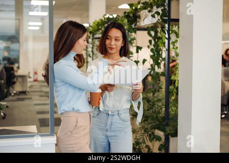 Ritratto di due colleghi multirazziali che chiacchierano in ufficio mentre bevevano un caffè e guardavano documenti cartacei. Condivisione femminile caucasica Foto Stock