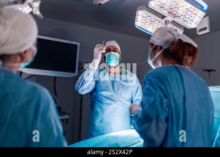 Un team medico di chirurghi vestiti di scrub blu esegue un intervento chirurgico sotto le luci accese, dimostrando una concentrazione approfondita e l'uso di avanzate Foto Stock