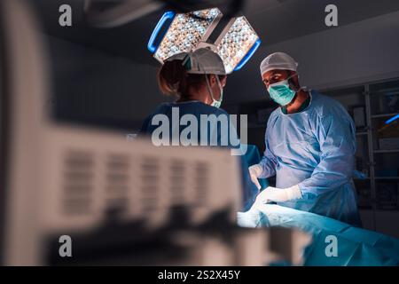 Sotto le luminose luci chirurgiche in sala operatoria, i chirurghi professionisti che indossano abiti e cappucci blu eseguono un'operazione con messa a fuoco e preci intensi Foto Stock