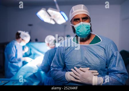 Un chirurgo maschile, vestito di scrub e una maschera chirurgica, si trova in sala operatoria con le mani sul petto, mentre il suo team chirurgico lavora su un paziente Foto Stock