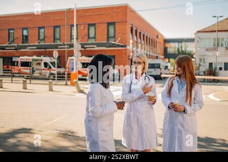 Tre medici donne in camice bianco si stanno godendo una pausa caffè fuori dall'ospedale, impegnandosi in una conversazione amichevole Foto Stock