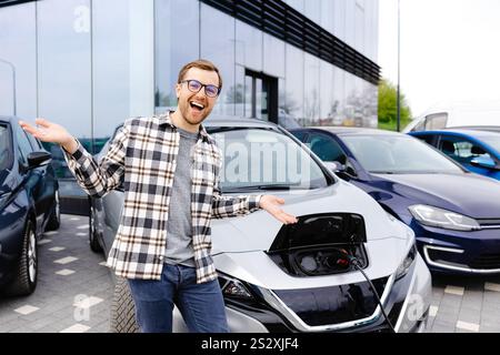 Un giovane uomo si trova accanto alla sua nuova auto elettrica e si riposa mentre la macchina è in carica. Foto Stock