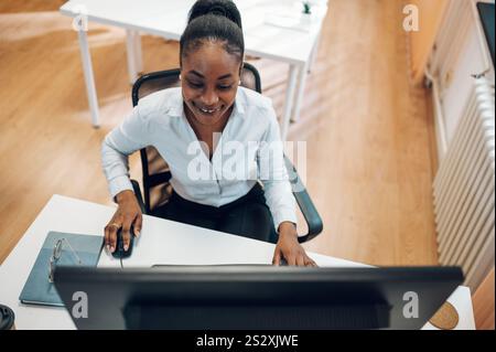 Concentrata giovane donna d'affari afroamericana che lavora su un notebook mentre è seduta al tavolo in ufficio. Esperto aziendale sicuro di sé. E-commerce Foto Stock