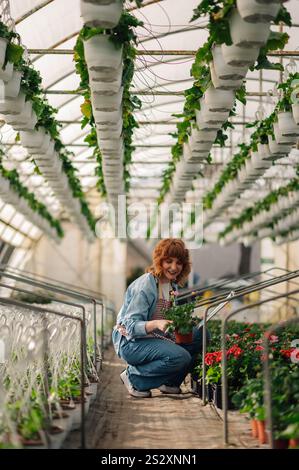 Ritratto di donna giardiniere sorridente allo zenzero accovacciato alla serra circondato da vasi di vegetazione e che tiene in mano vasi di fiori. Felice pianta nur Foto Stock