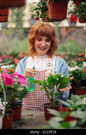 Giardiniere donna che utilizza uno spruzzatore d'acqua per innaffiare gli alberelli colorati mentre si trova in un grande locale floreale con molte pentole sui tavoli e appese al c Foto Stock