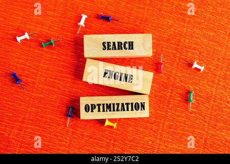 Tre blocchi di legno etichettati ricerca, motore e ottimizzazione sono visualizzati in modo creativo tra i pulsanti colorati su una superficie arancione vivace, evidenziata Foto Stock