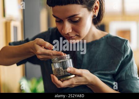 Ritratto di una giovane donna che sorride amichevole mentre tiene in mano un barattolo con le erbe che sta crescendo nel suo giardino di casa in salotto. Prendersi cura delle piante domestiche e.. Foto Stock