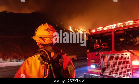 Malibu, California, Stati Uniti 7 gennaio 2025. Un Vigili del fuoco di LA County in un cappello da 89 è acceso rosso e giallo dalle luci dei veicoli di emergenza. Un incendio scoppiò il 7 gennaio 2025, come si vede dalla PCH (Pacific Coast Highway) nella ricca città di Pacific Palisades, ai margini di Malibu, CA, a causa di venti di Santa Ana insolitamente forti che superavano i 100 MPH (Credit Image: © Amy Katz/ZUMA Press Wire) SOLO PER L'USO EDITORIALE! Non per USO commerciale! Foto Stock