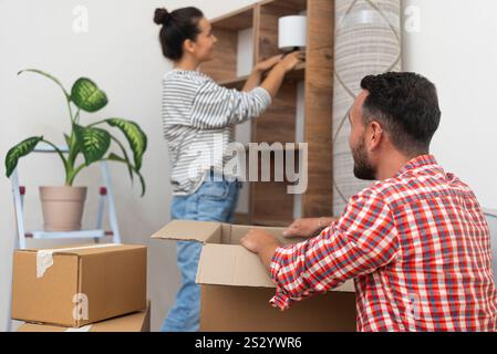 la giovane famiglia felice si trasferisce nella nuova casa, disimballando e adornando uno scaffale vuoto con una lampada Foto Stock