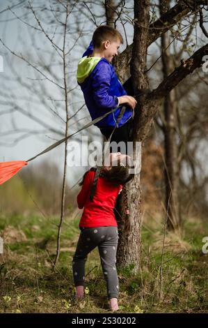 Due bambini si dedicano a divertenti attività all'aperto. Un ragazzo sale su un albero, assistito da una ragazza sotto. La scena è ambientata in un ambiente naturale con alberi A. Foto Stock